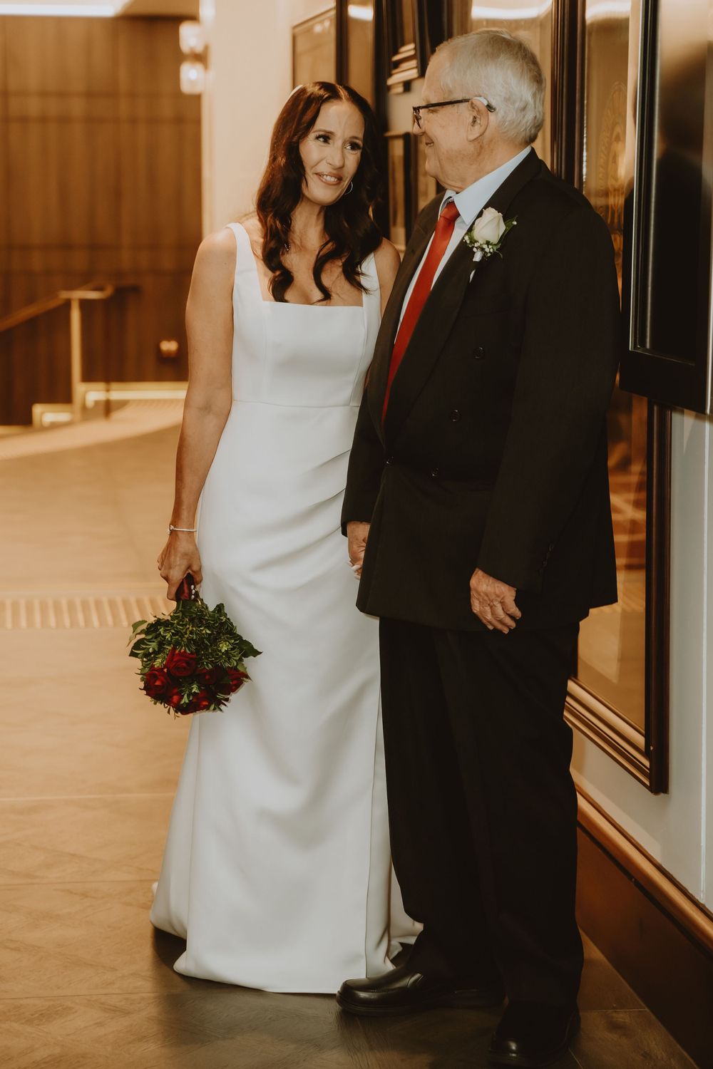 A bride in white dress walks alongside an elegantly dressed older gentleman in a dark suit through a hallway setting.