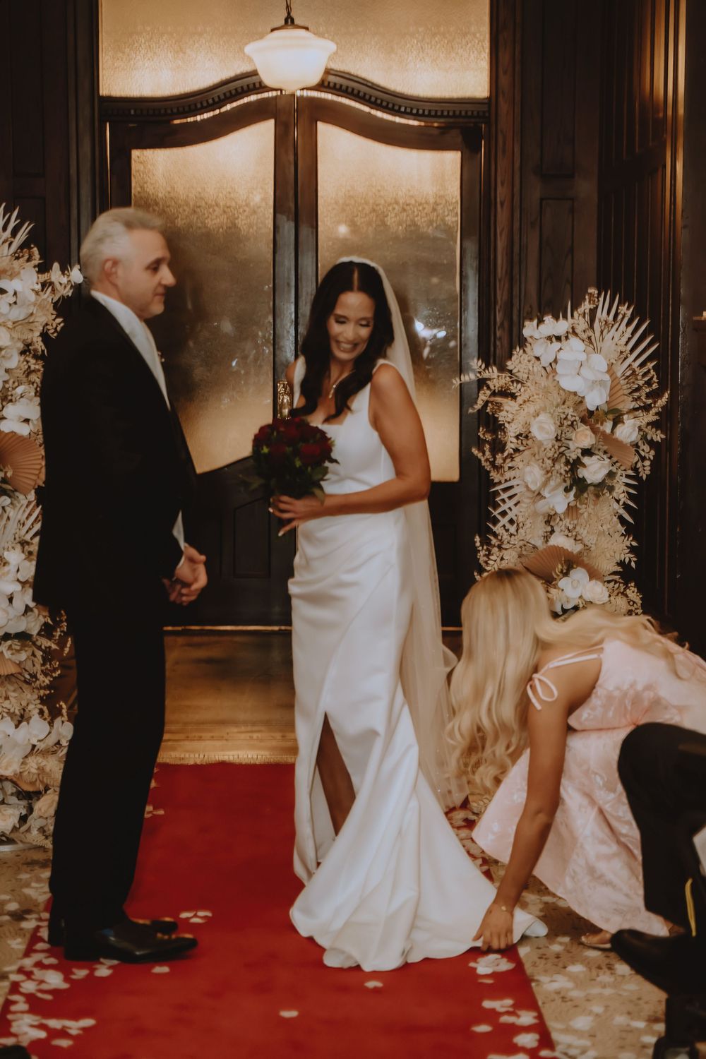 Wedding couple dancing and celebrating with guests on luxurious red carpet surrounded by elegant white floral arrangements.