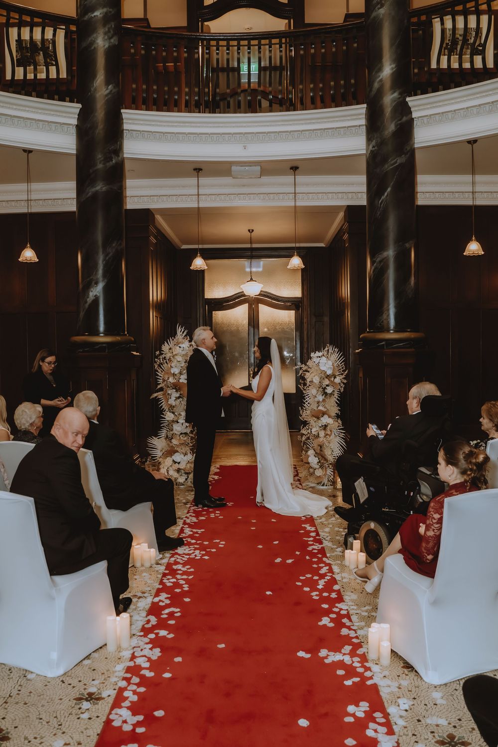 Wedding ceremony setup with red carpet aisle, white chair covers, and elegant floral arrangements in an indoor venue.
