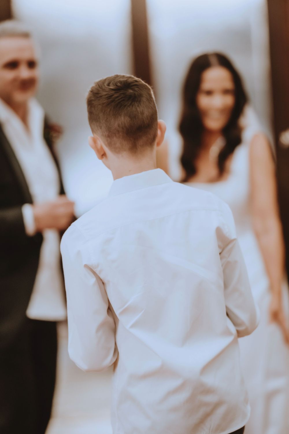 A child in white clothing walks down a hallway as blurred figures stand in the background.