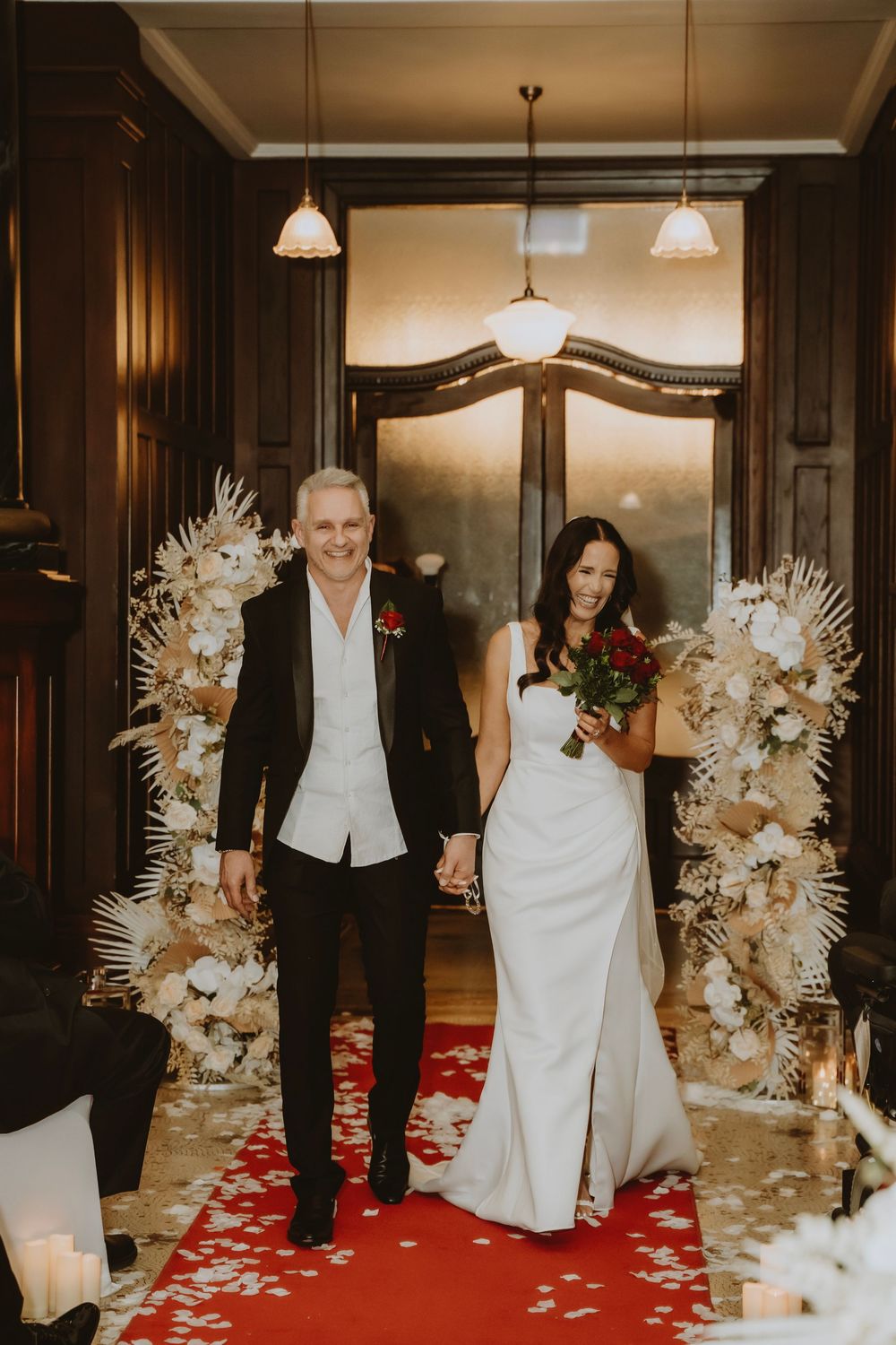 A couple walks down a red carpet aisle between elegant white floral arrangements in a warmly lit indoor venue.