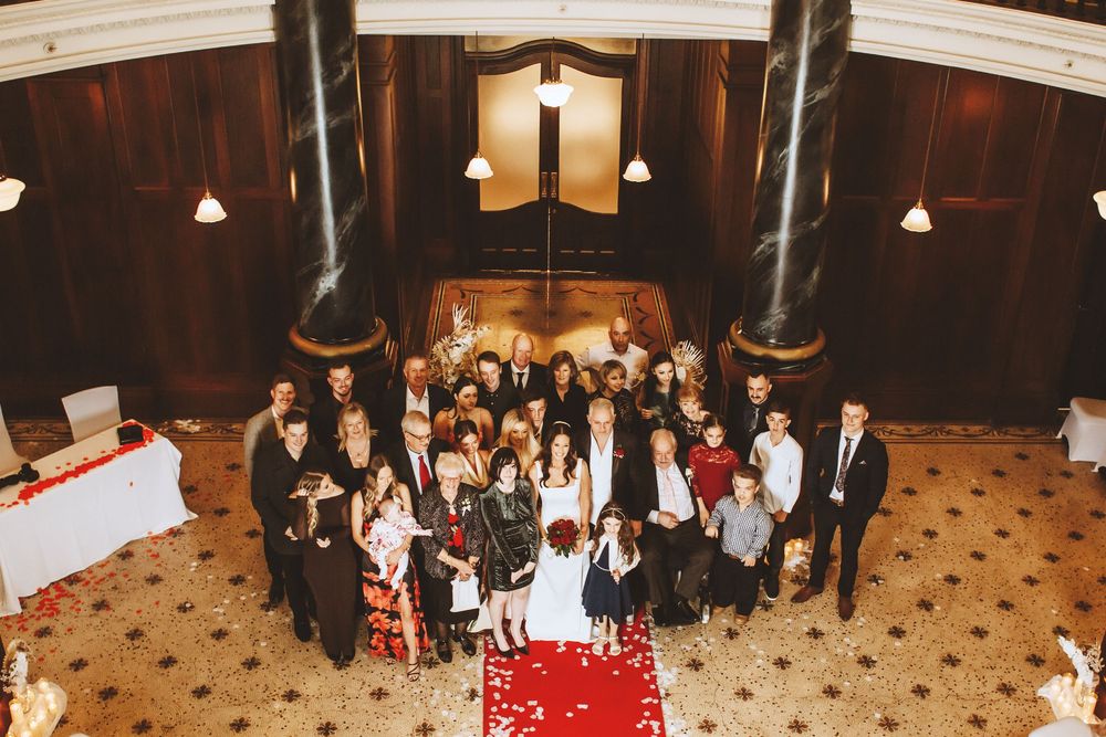 Wedding guests gathered together in formal attire on red carpet for group photo in elegant wood-paneled hall.