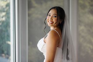A bride in a white wedding dress and veil poses by a bright window on her special day.