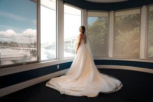 A flowing white wedding gown with a long train is captured in front of bright bay windows overlooking a marina.