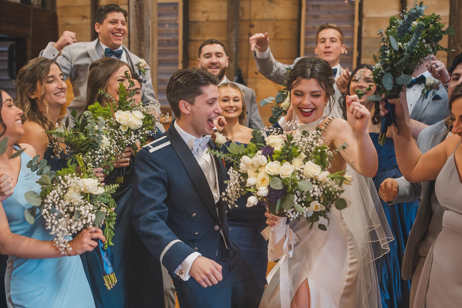 Wedding party celebrating with joy in rustic barn setting with floral bouquets.