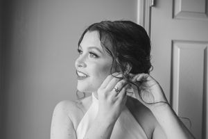 A woman in black and white puts on earrings while getting ready, showing a joyful expression.
