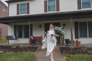 Person in white dress stands in front of traditional white house with covered porch.