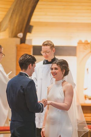 A couple exchanges rings during a wedding ceremony in a church while officiant stands behind them in traditional attire.