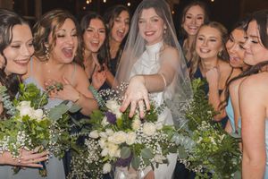 A group of bridesmaids in gray dresses gather around a bride in white, all laughing and showing off their bouquets.