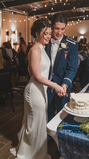 A couple in formal wedding attire cutting their white wedding cake together at an indoor reception.