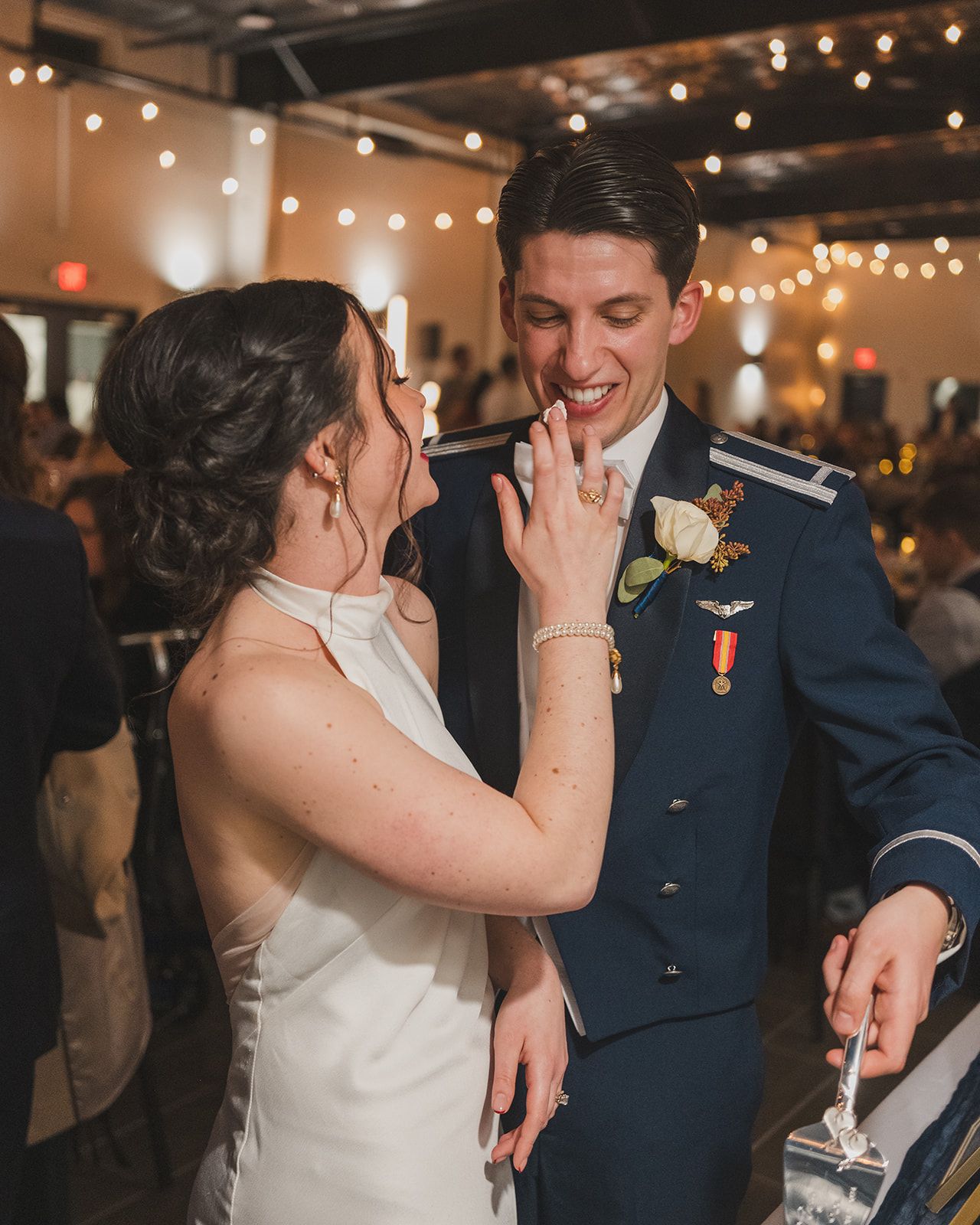 A couple in wedding attire share a joyful moment feeding each other cake at their reception with string lights in background.