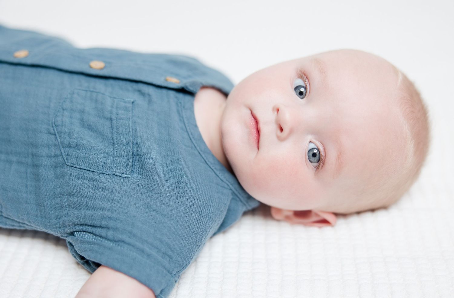 Baby wearing blue linen outfit lays on white blanket looking up with bright eyes.