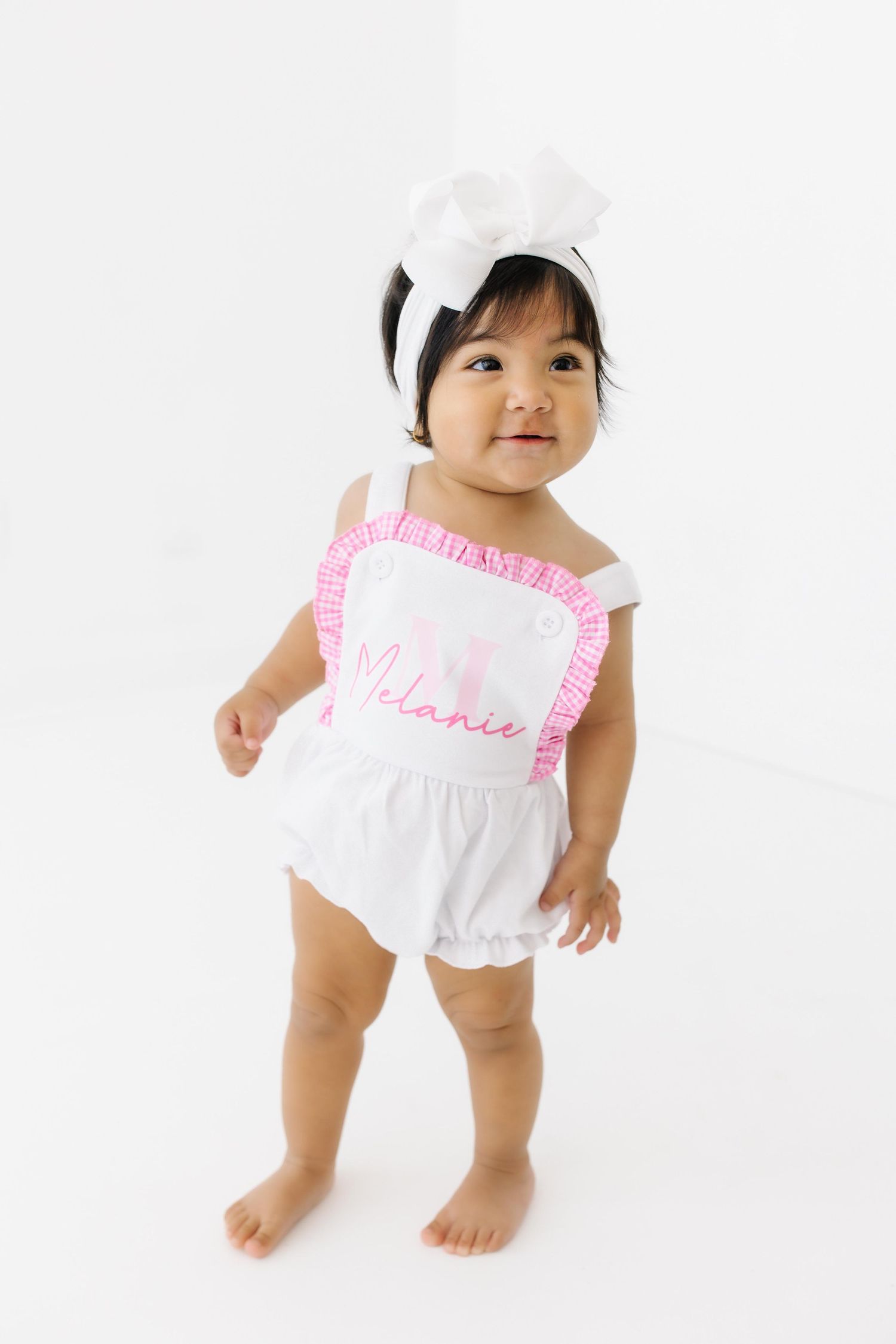 Baby wearing white ruffled sunsuit and cap with pink trim against white background.