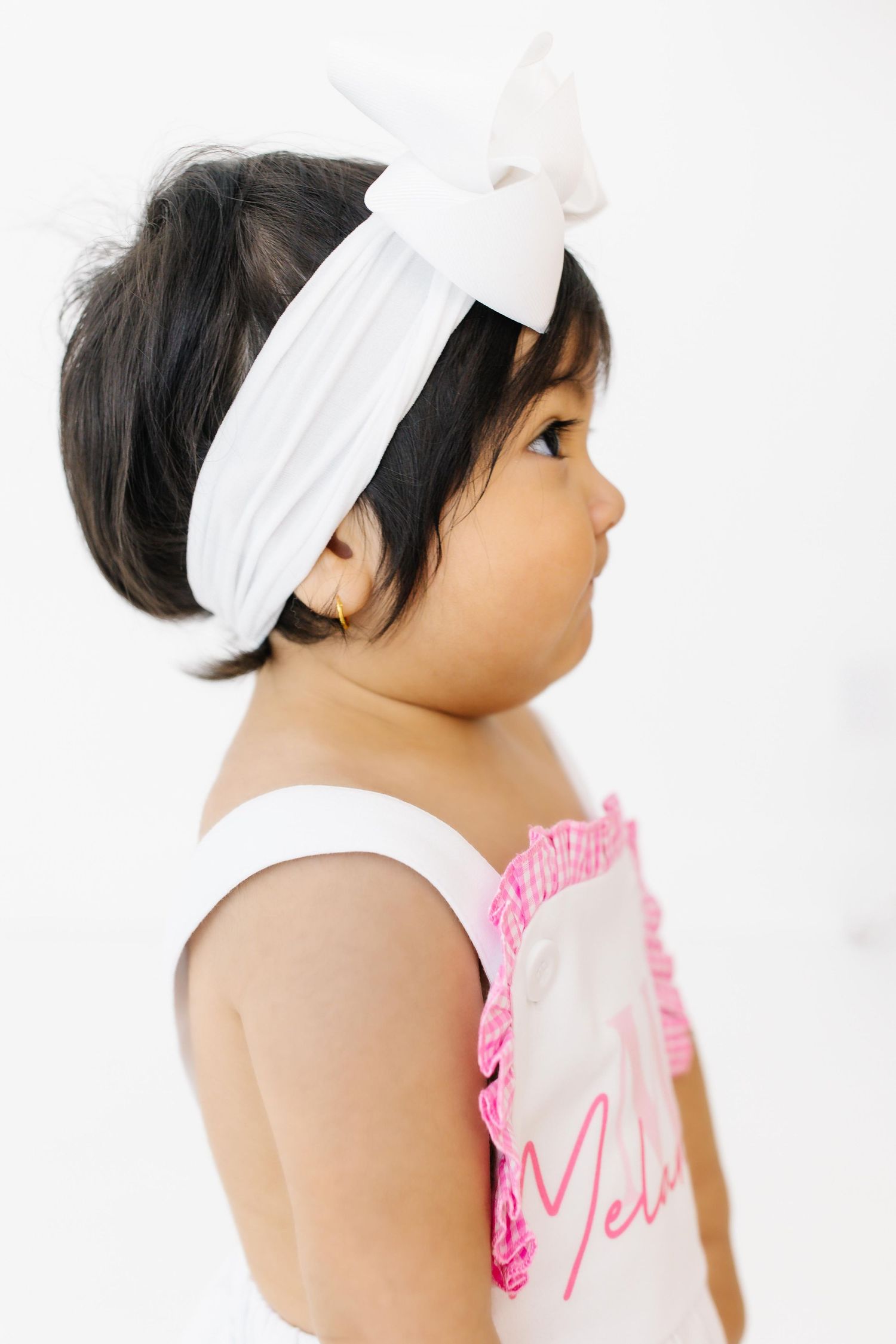 Side profile of a baby wearing a white headband and pink ruffled clothing against a light background.