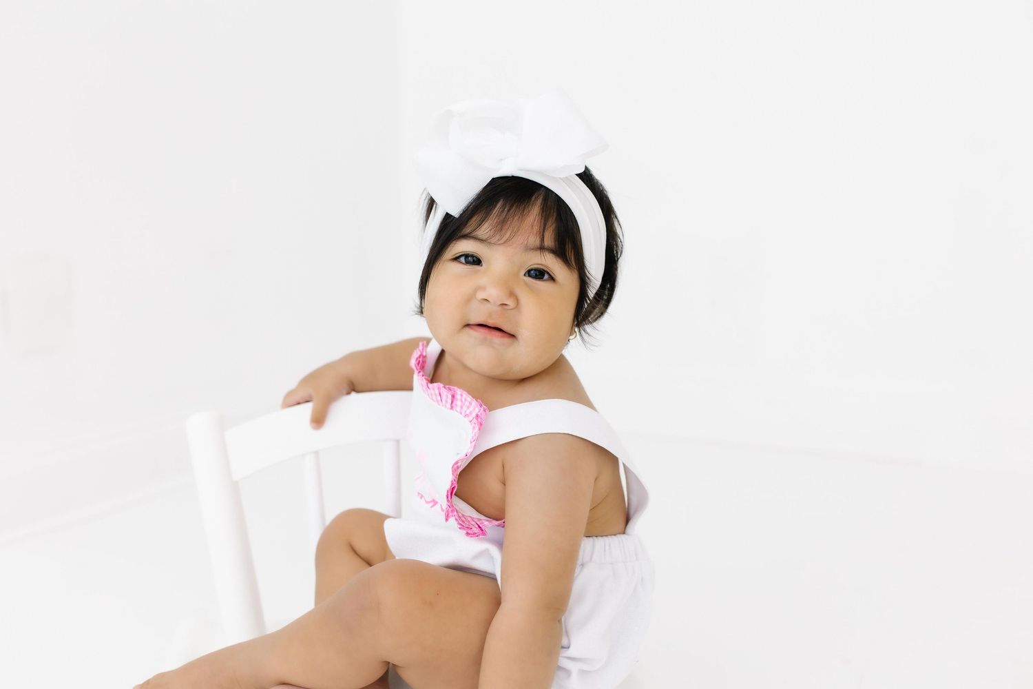 A small child wearing a white headband and sleeveless top sits on a white chair against a plain background.