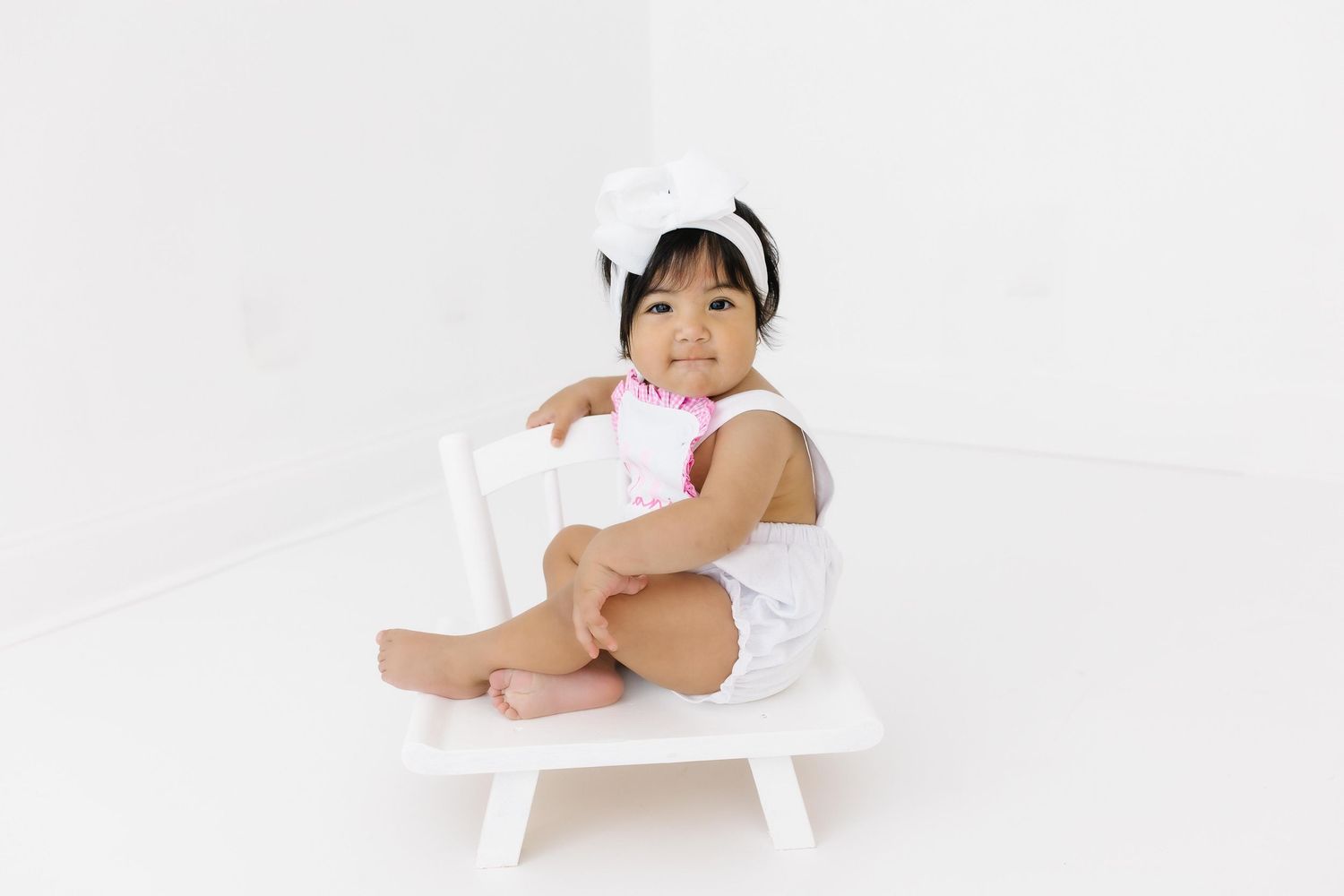 Small child sitting on a white wooden chair wearing white outfit and pink accessory against white background.