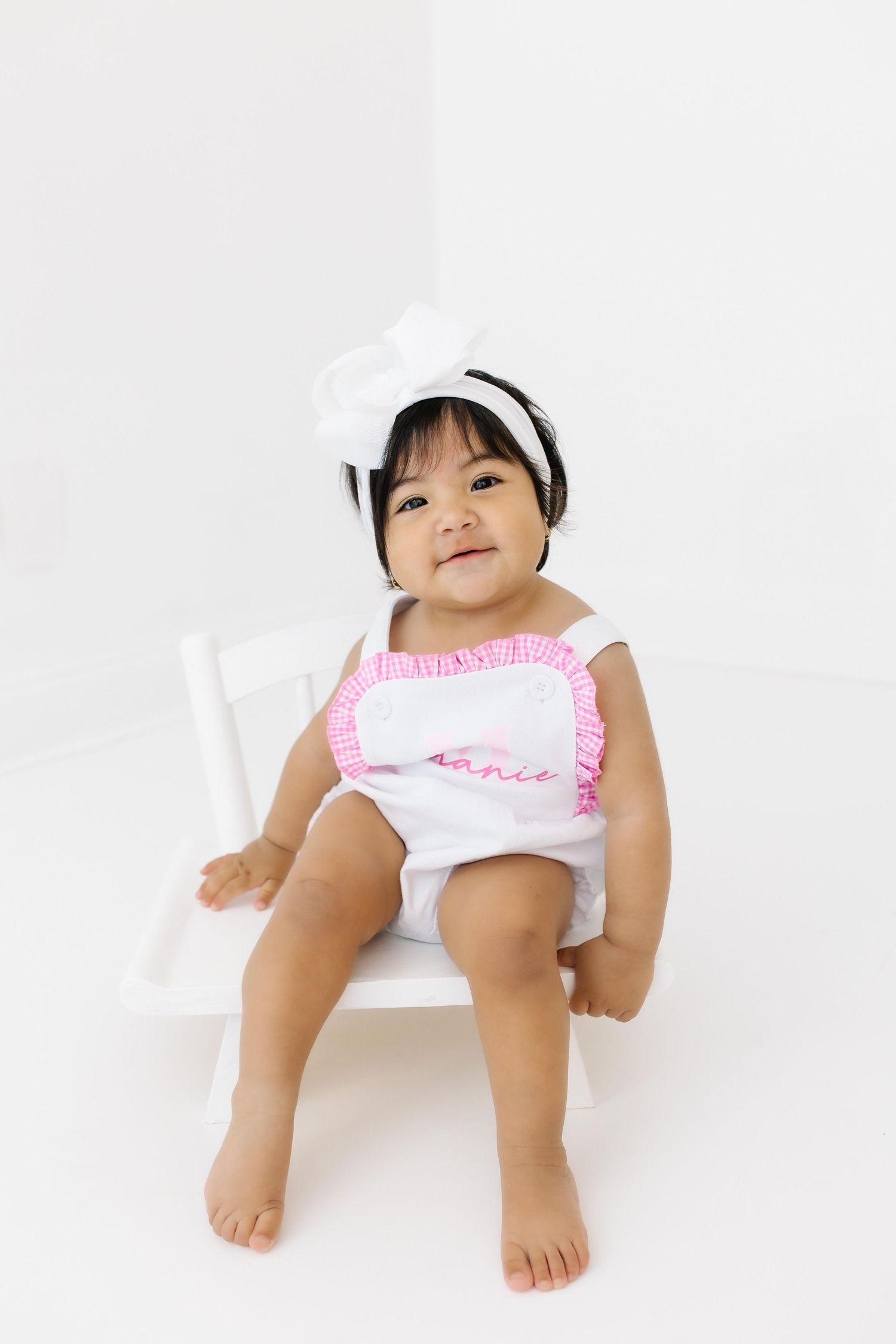 A toddler wearing a white and pink ruffled outfit sits on a white chair against a plain white background.