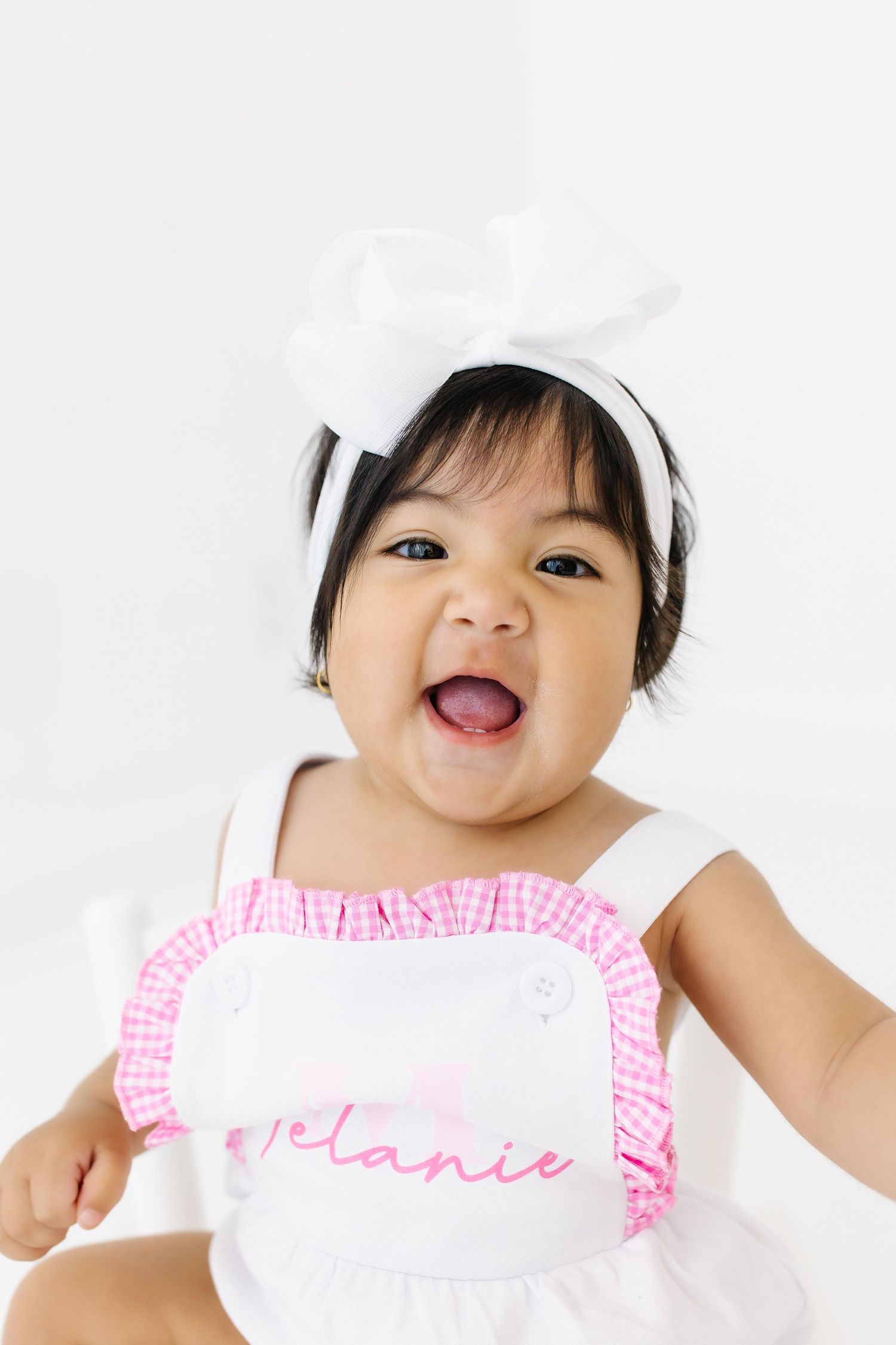 Baby wearing pink and white ruffled dress shows joyful expression while lying on white background.