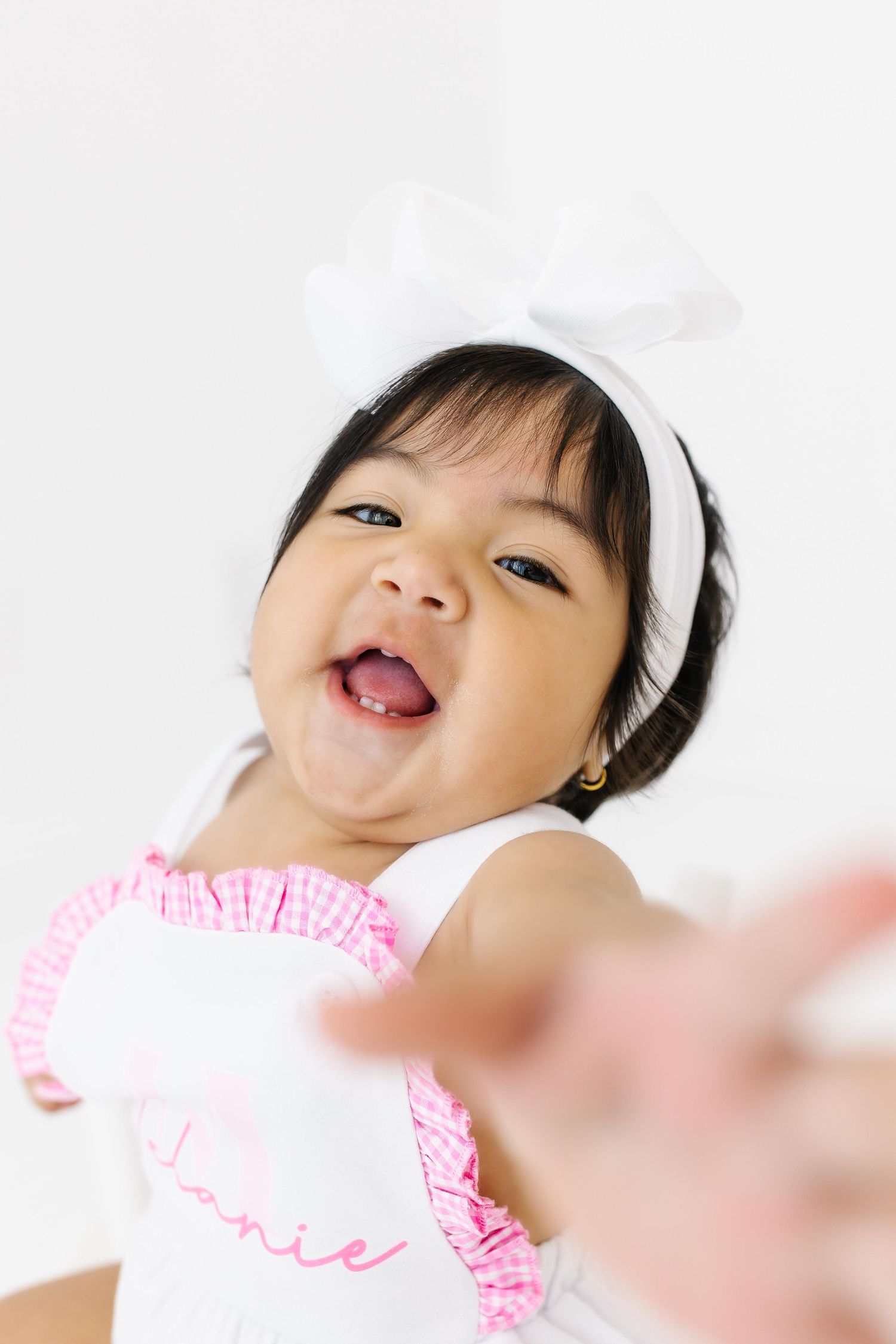 Young child wearing a white dress with pink ruffles smiles joyfully while lying down on a bright background.