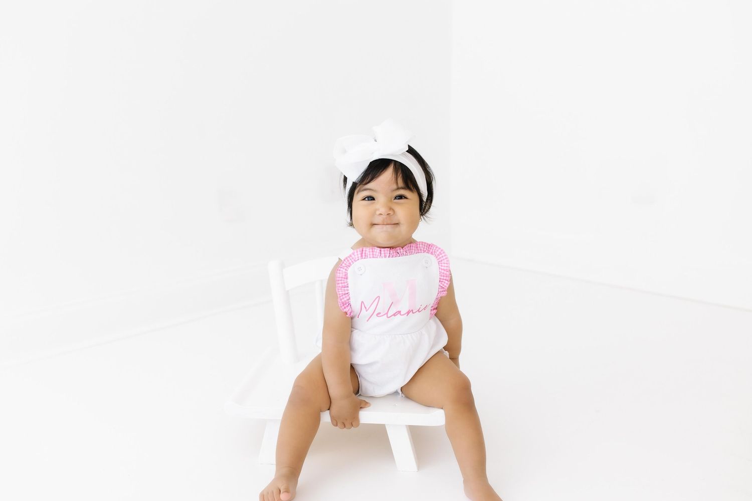 Baby wearing a pink and white outfit sits on a small white stool against a white background.