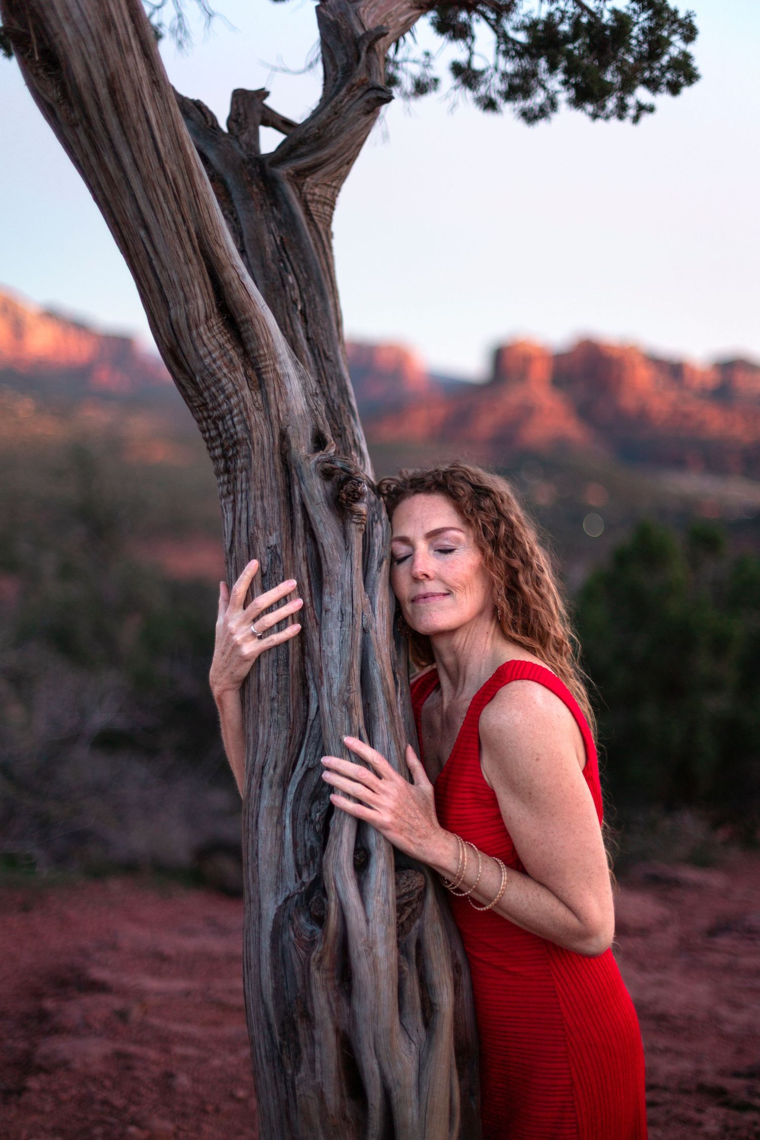 Woman in red dress embracing juniper tree with eyes closed near Sedona red rocks at dusk.