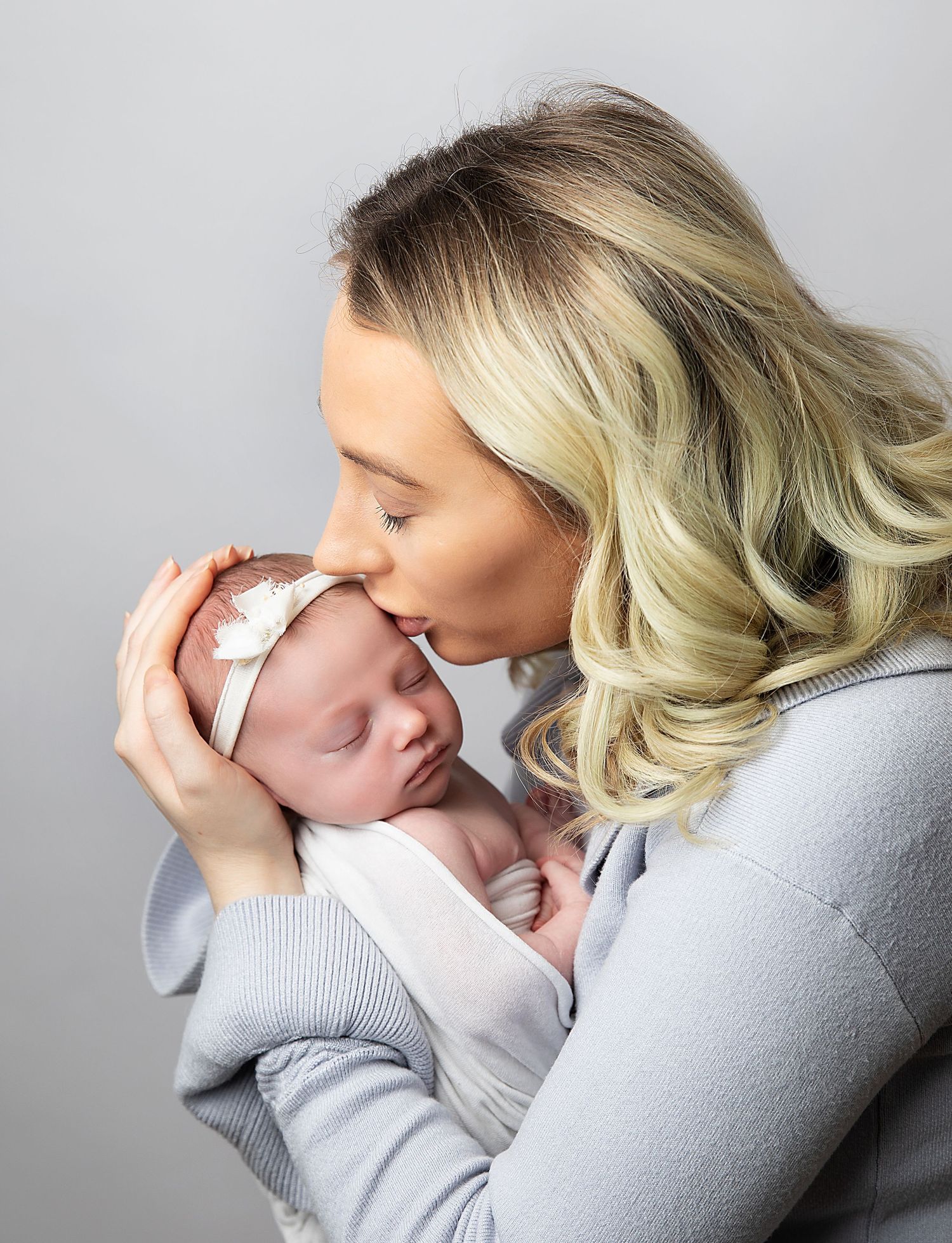 A tender moment shows a woman in a gray sweater gently kissing a sleeping newborn baby wrapped in white.