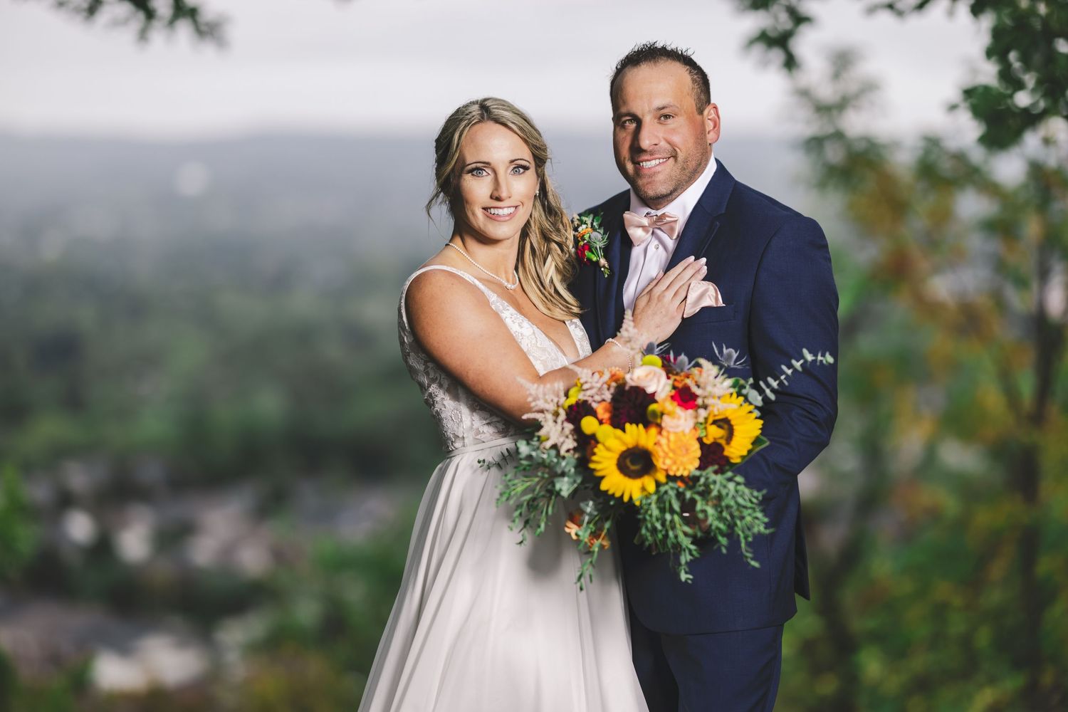 A newlywed couple poses together outdoors with a vibrant sunflower bouquet against a misty mountain backdrop.