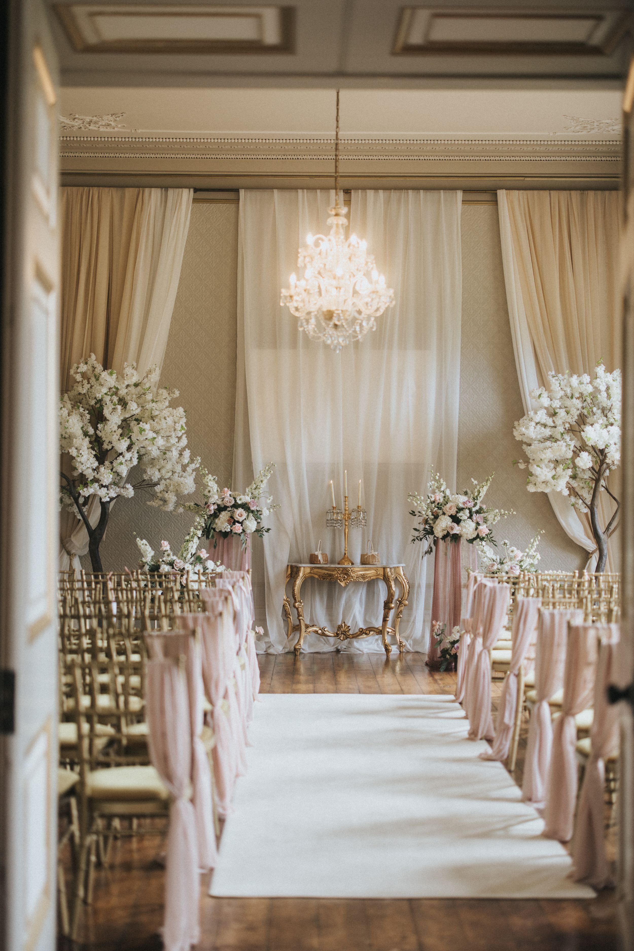 Elegant wedding ceremony aisle lined with blush pink fabric and tall white floral arrangements under crystal chandeliers at Rise Hall.