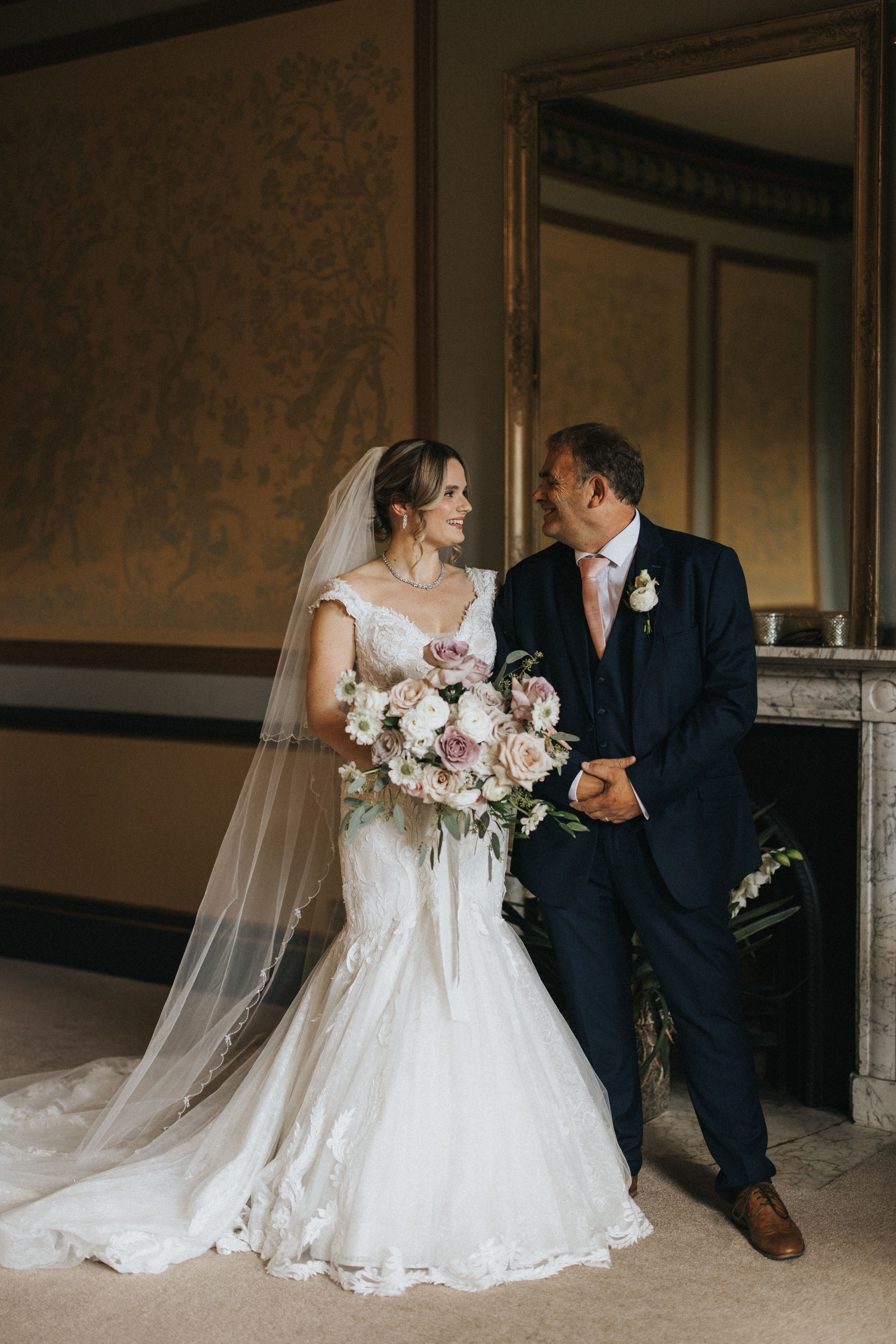 A couple in formal wedding attire poses in an elegant interior space with a large ornate mirror and vintage wallpaper at Rise Hall.