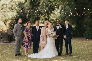 A wedding party poses together outdoors with string lights and greenery in the background at Rise Hall.