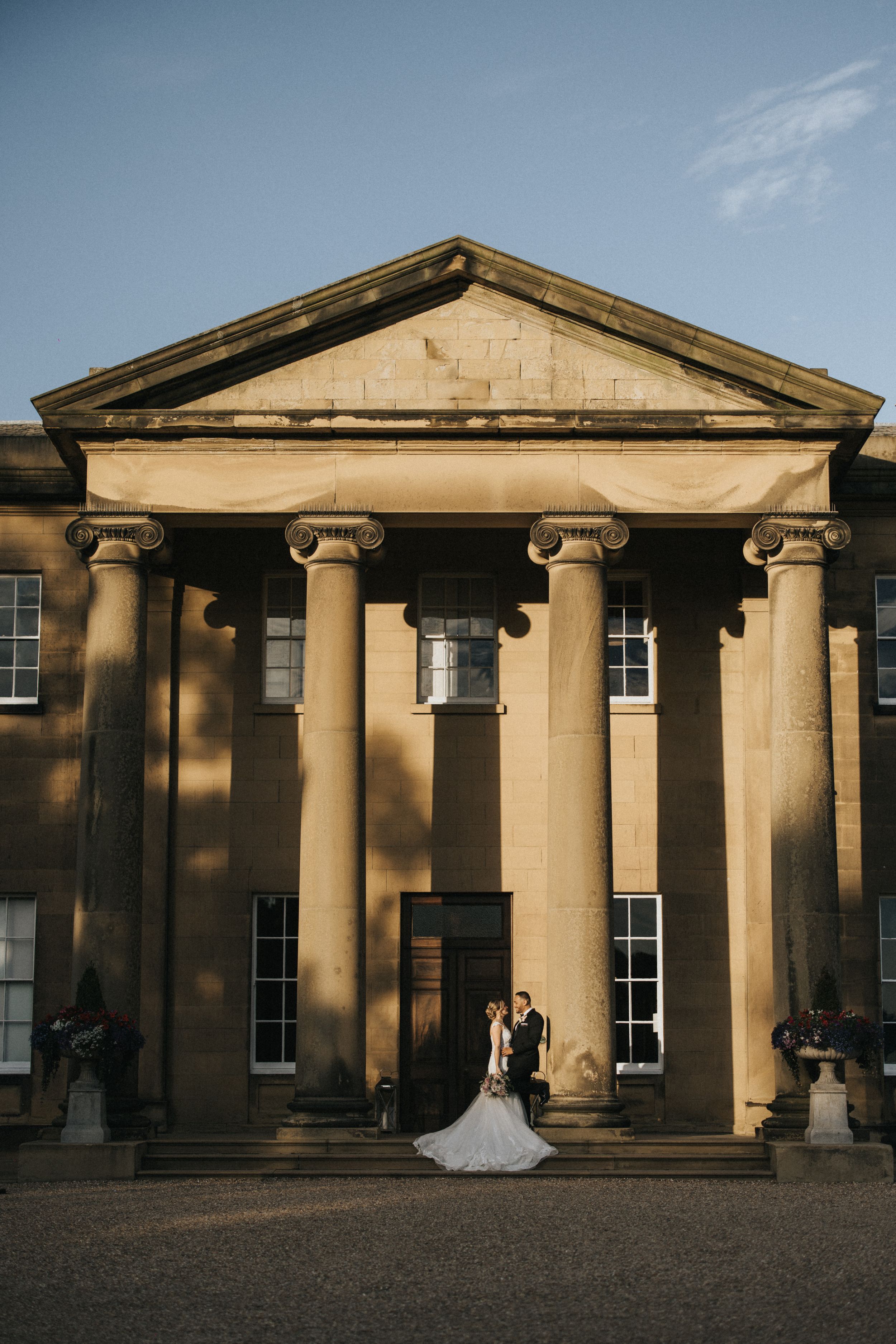 Elegant couple standing in front of a grand neoclassical building at Rise Hall with towering stone columns and triangular pediment.