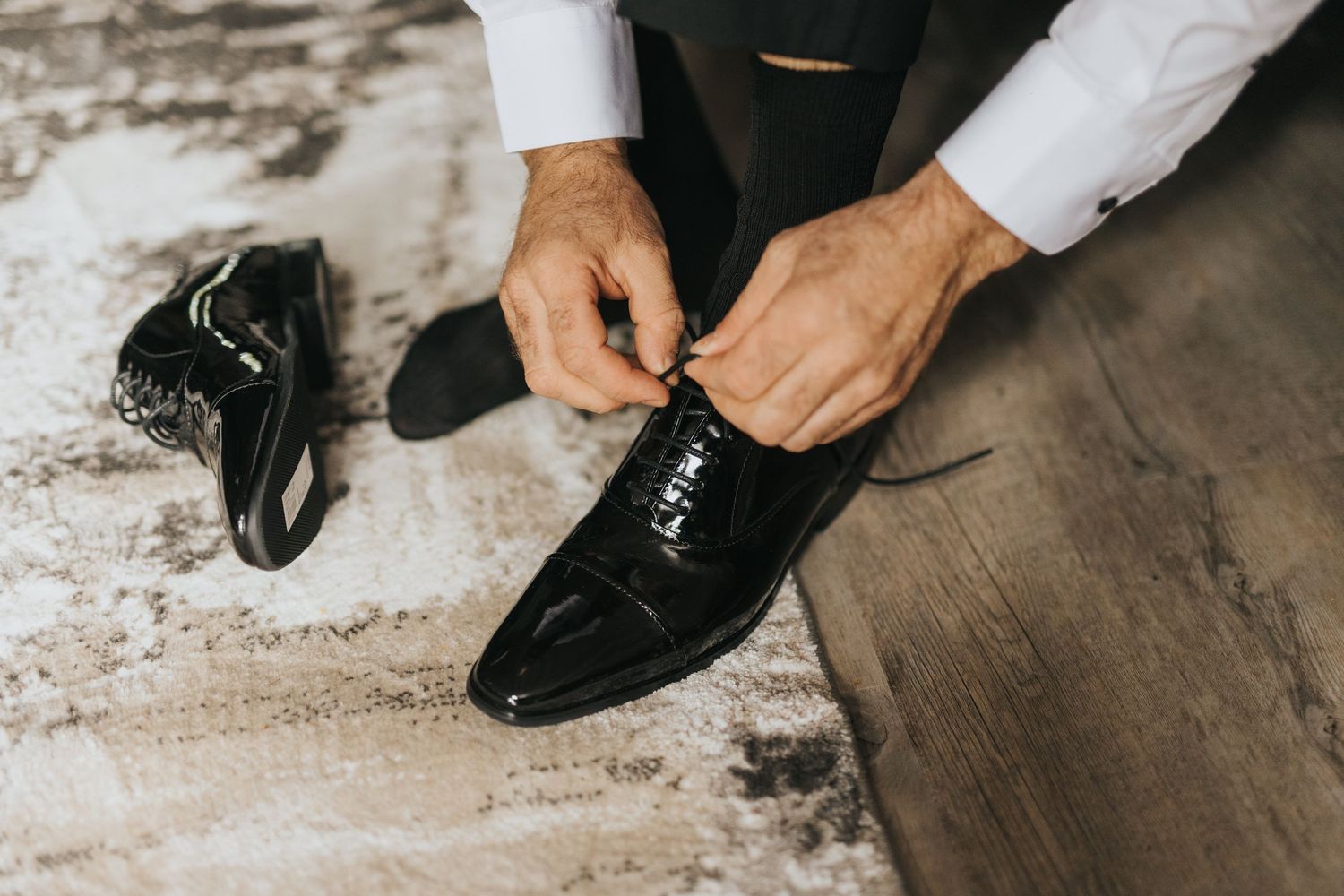 Groom tying laces on black leather dress shoes on a wooden floor with scattered white powder or dust in the background.