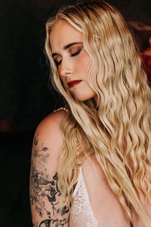 Close-up portrait showing wavy blonde hair, floral arm tattoo and lace detail in moody lighting. Nice & Naughty Store Event Grand Rapids MI