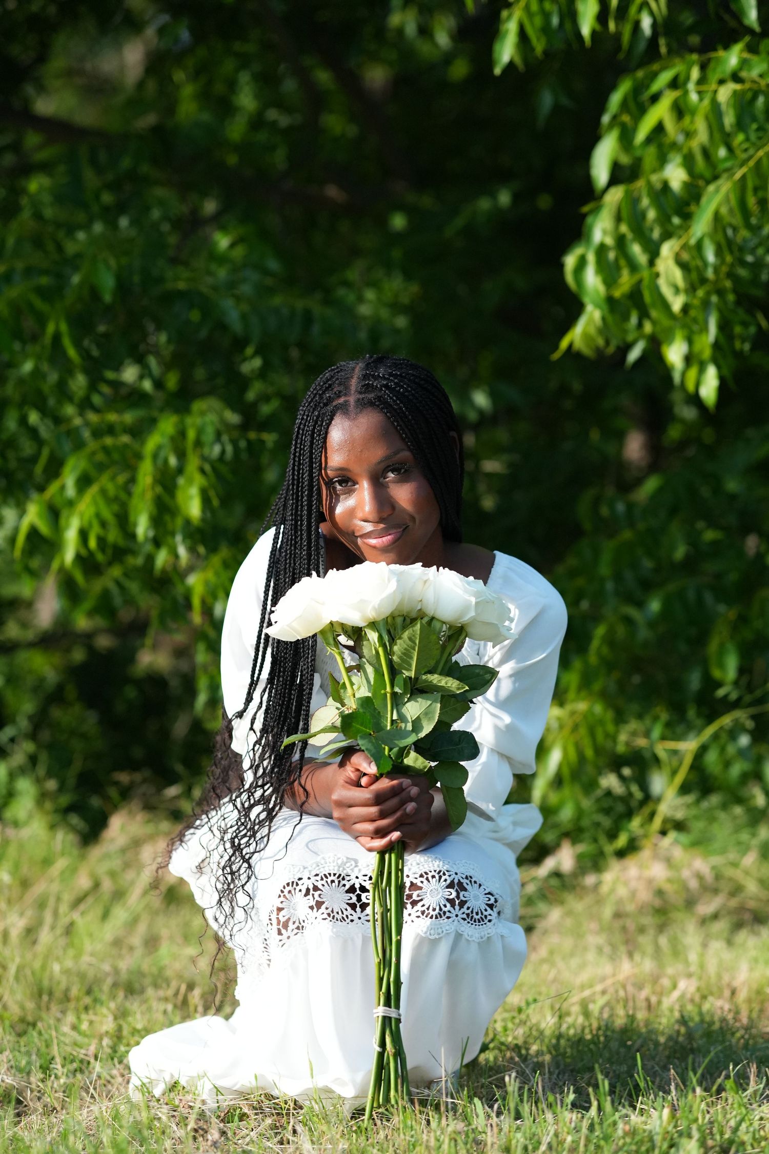 A person in a white dress kneels on grass holding white roses against a backdrop of lush green foliage.