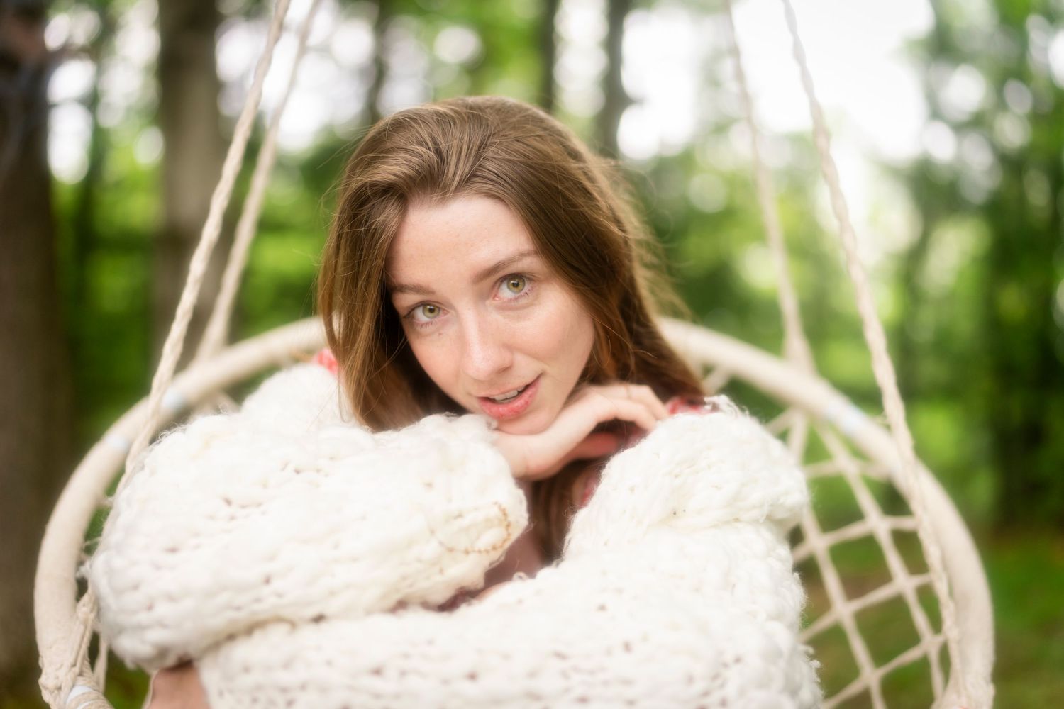 Person relaxing in macrame hammock chair wearing cozy white knit sweater in outdoor forest setting with soft bokeh light.