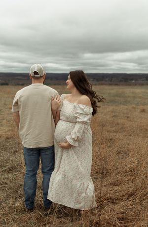 A couple stands together in a brown field under moody cloudy skies, with her wearing a white off-shoulder dress.