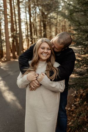 A couple shares intimate moments in a sunlit forest setting, wearing casual sweaters and embracing playfully.