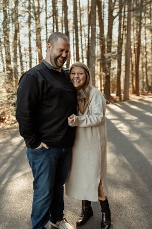 A couple laughs together on a sunlit forest path during an autumn engagement photo session.