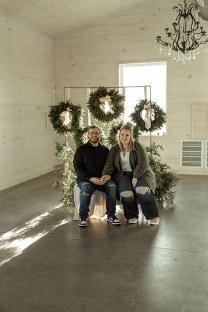 A couple sits together on a concrete floor in a bright white room decorated with hanging wreaths and chandeliers.