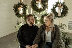 A couple sits together in front of three evergreen Christmas wreaths hanging on a white wall.