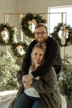 A couple embraces in front of illuminated holiday wreaths hanging on a white wall in warm lighting.