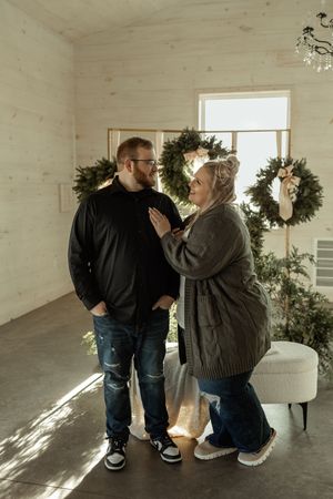 Two people share a tender moment in a sunlit room with white walls and evergreen wreaths hanging as decor.
