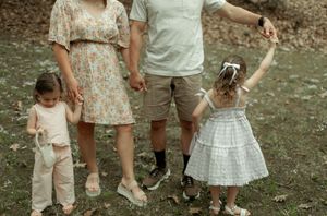 A casual family portrait showing parents and two young children holding hands in an outdoor setting.