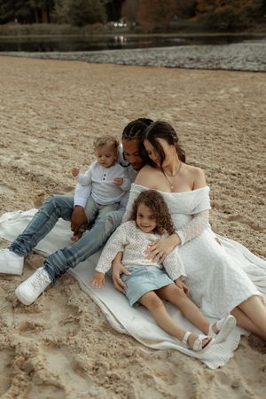 Family relaxing together on a sandy beach blanket in casual white and denim outfits.