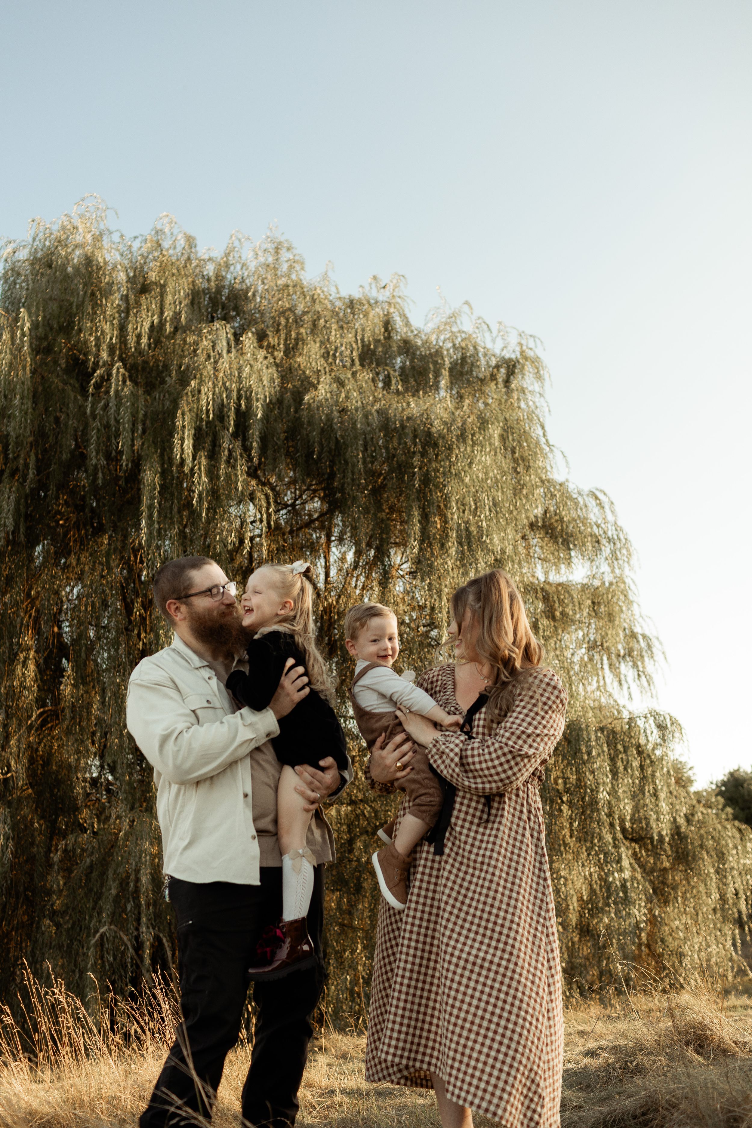 Family shares playful moments together under a weeping willow tree in warm evening light.
