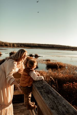 Two people stand on a wooden boardwalk overlooking a scenic marsh landscape at sunset in warm golden light.