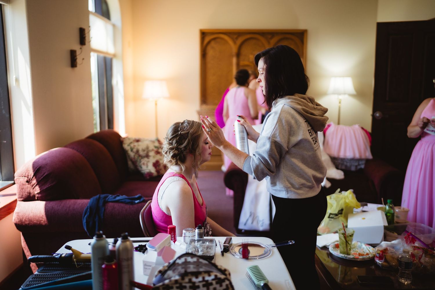 Wedding party members in pink dresses get ready in hotel room with natural lighting and personal touches evident.