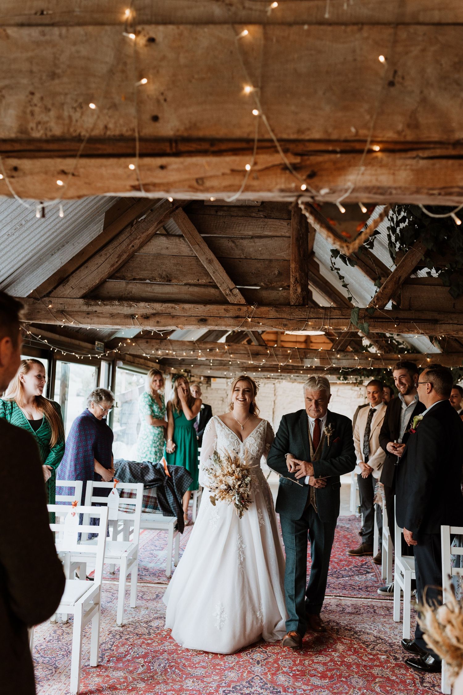 A rustic barn wedding ceremony setup with string lights overhead, white chairs, and an ornate red patterned carpet down the aisle.