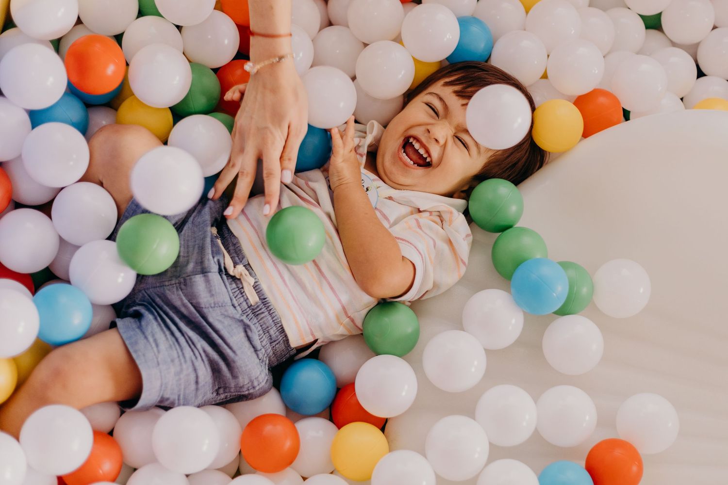 Child playing and laughing in a colorful ball pit filled with white, orange, blue and green plastic balls.