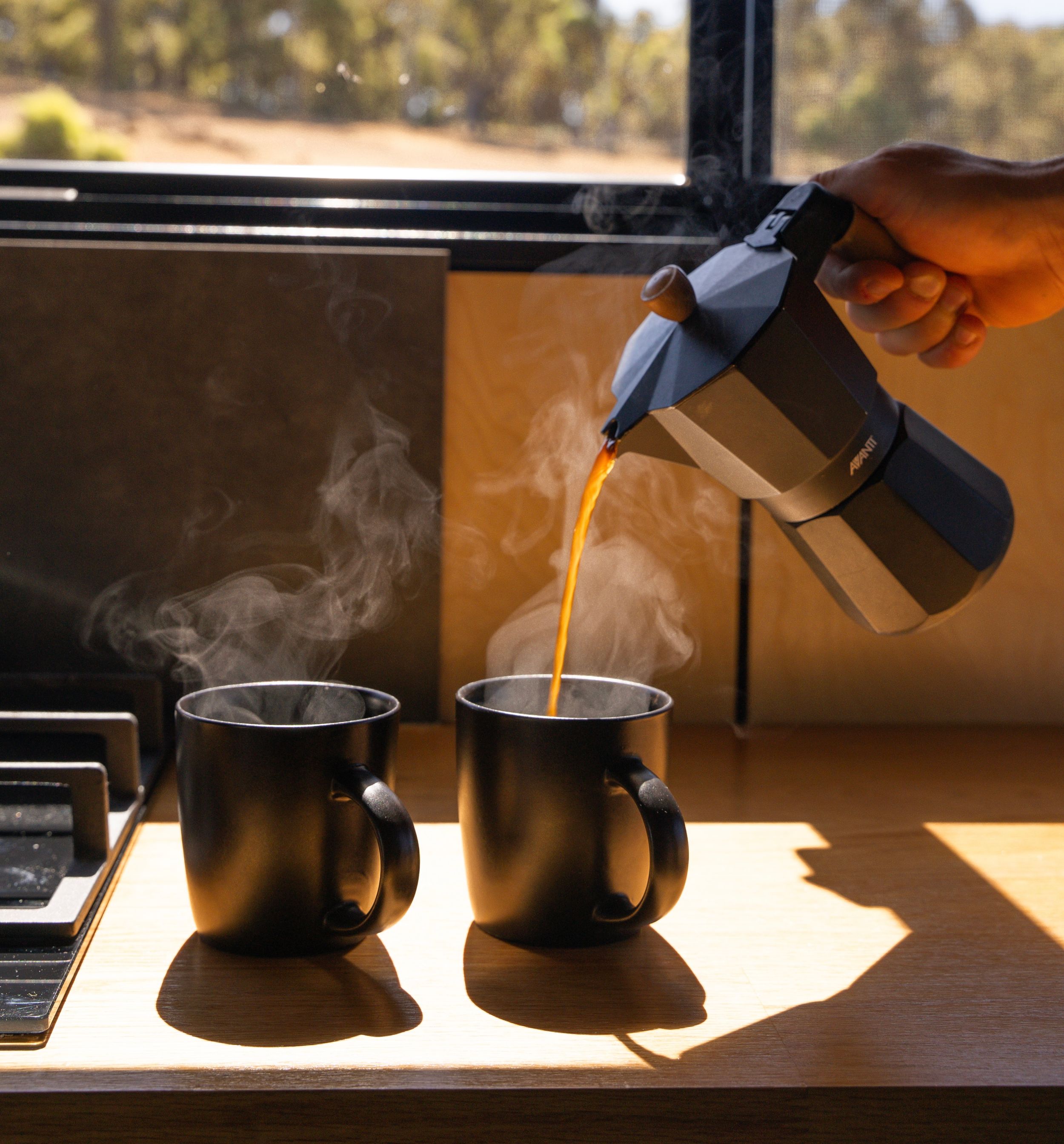 Steaming coffee being poured from a moka pot into two dark mugs on a wooden surface in morning sunlight.